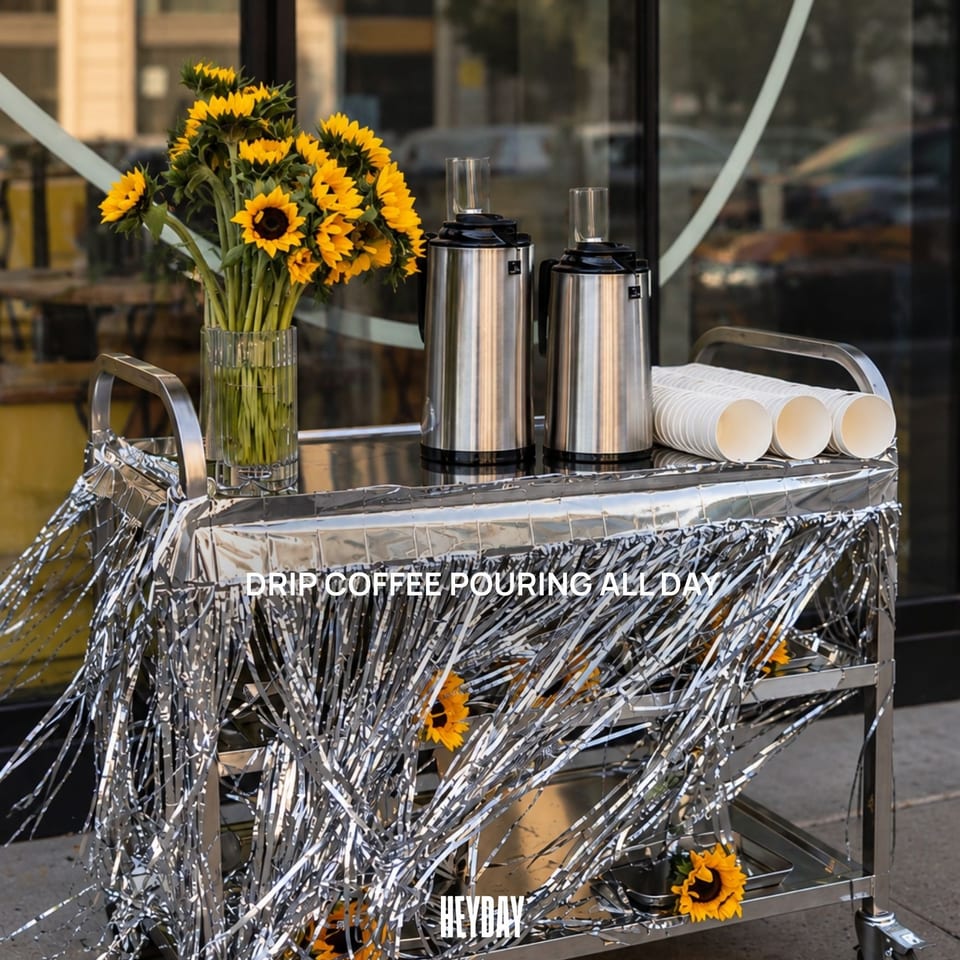 Coffee service cart decorated with sunflowers, silver tinsel, stainless-steel carafes, and stacked cups outside Heyday café.