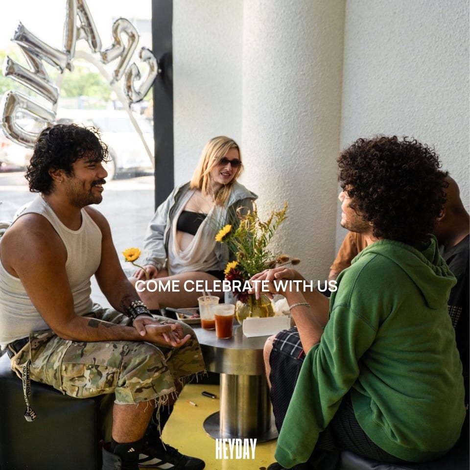 Friends gather around a restaurant table with drinks and flowers beneath a celebratory Heyday message.