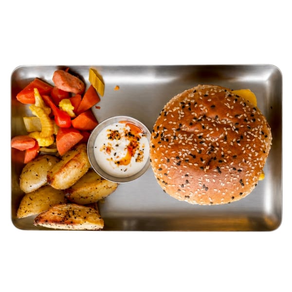 Top-down view of a sesame seed burger, roasted potatoes, mixed vegetables, and dipping sauce on a metal tray.