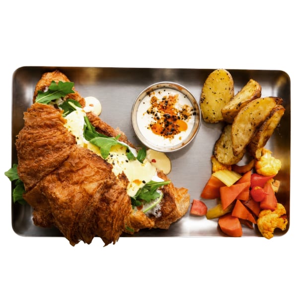 A croissant sandwich with arugula, cheese, and sauce, served with potato wedges, yogurt, and pickled vegetables.